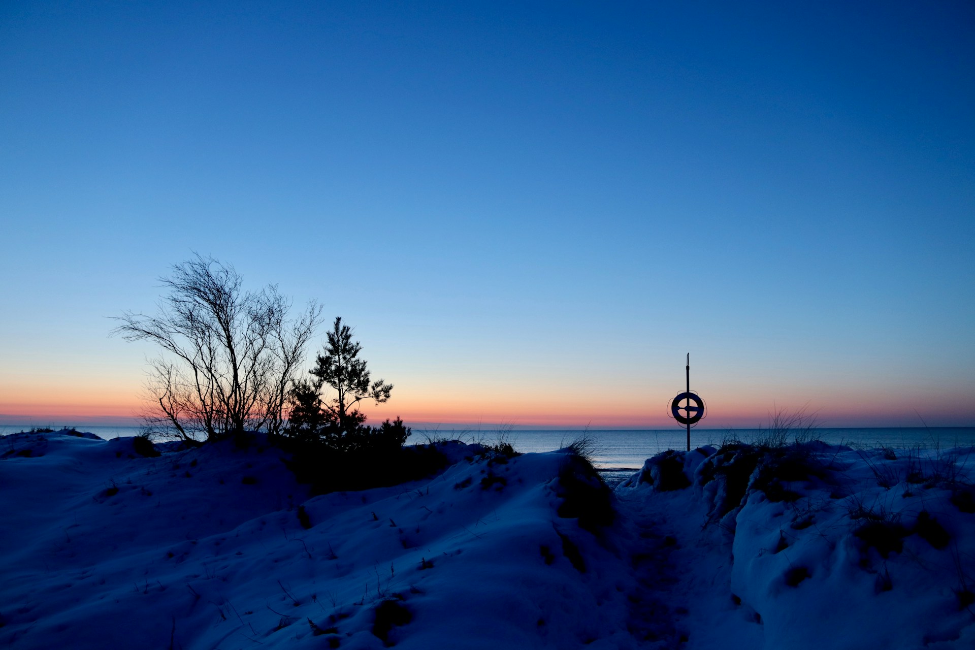 Solnedgang over et snedækket strandlandskab med sparsomme, bladløse træer, en fjern vandmasse og en forfatters signaturkorsformede livredderstand.